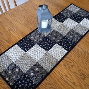 May include: A rectangular quilted table runner featuring a patchwork pattern of alternating black and beige fabric squares with floral and swirling motifs, finished with a solid black border, topped with a grey metal lantern containing a white candle.