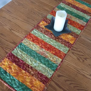 May include: A rectangular quilted table runner featuring alternating fabric strips in shades of deep red, forest green, and golden yellow with floral and leaf patterns. A white pillar candle sits in a black star-shaped metal candle holder.