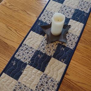 May include: A quilted table runner with a patchwork design in navy blue and beige floral patterns. A white candle sits on a star-shaped metal holder. The runner is on a wooden table.