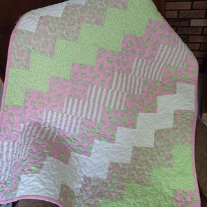 May include: A pink and green baby quilt with a chevron pattern. The quilt is made of different fabrics with various patterns, including polka dots, stripes, and floral designs.