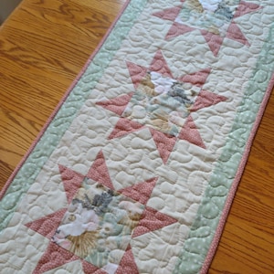 May include: A table runner with a white background and three pink stars with a floral pattern. The stars are outlined in pink and the runner has a green border with white polka dots.