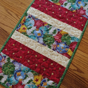 May include: A quilted Christmas table runner with a green border. The runner features alternating red dotted and beige striped sections. The beige stripes have a gold snowflake pattern. The red sections have a dotted pattern. The center section has a pattern of colorful Christmas ornaments.