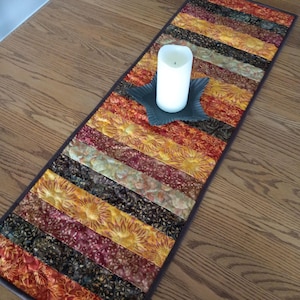 May include: A rectangular fabric table runner featuring a patchwork design of horizontal strips in shades of orange, yellow, brown, and burgundy with floral patterns. A white pillar candle sits on a dark star-shaped metal base in the center.