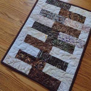 May include: A brown and white patchwork quilt with a geometric pattern. The quilt is made of various fabrics with different patterns and colors, including brown, white, and blue. The quilt is edged with a brown border.