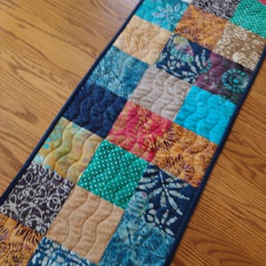 May include: A colorful patchwork table runner with a dark blue border. The runner is made of various patterned squares in shades of blue, gold, green, and red. The squares have quilted details, creating a textured surface. The runner is placed on a wooden table.