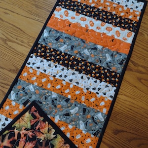 May include: A rectangular quilted table runner featuring alternating diagonal fabric strips with Halloween patterns including orange pumpkins, white ghosts, black cats, and candy corn, finished with a solid black fabric border trim.