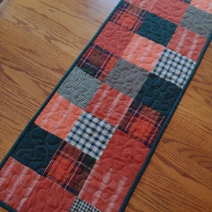 May include: A quilted table runner with a patchwork design. The runner features squares of plaid and gingham fabrics in shades of orange, red, navy, and gray. The edges are trimmed with a dark teal binding. The runner is placed on a wooden table.