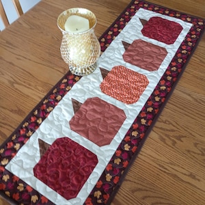 May include: A rectangular quilted table runner featuring five fabric pumpkins in shades of red, orange, and brown, set against a cream background with a dark border patterned with small autumn leaves, accompanied by a gold mosaic glass candle holder.