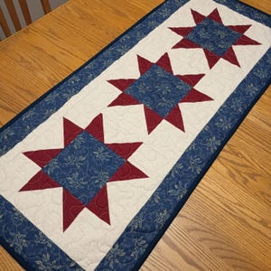 May include: A rectangular quilted table runner featuring three eight-pointed stars in a red and blue floral pattern against a cream background. The runner is bordered by a dark blue fabric with a repeating white vine and leaf print design.