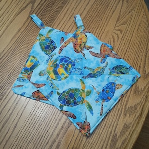 May include: Two square quilted pot holders featuring a vibrant pattern of multi-colored sea turtles in shades of blue, green, orange, and yellow against a light blue background. Each pot holder includes a small fabric loop for hanging storage.