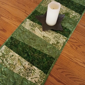 May include: A green and white quilted table runner with a star-shaped candle holder and a white pillar candle. The runner features various shades of green fabric in a striped pattern. The candle holder is dark brown.