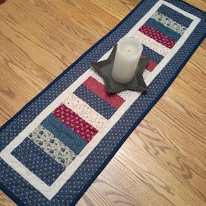 May include: A rectangular quilted table runner featuring a patchwork design of blue, red, and cream fabric strips with floral and dotted patterns. A dark gray star-shaped candle holder sits on the runner, holding a white pillar candle.