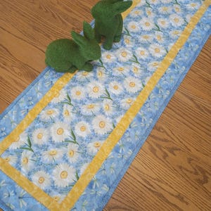 May include: A blue and white floral table runner with a yellow border, featuring a daisy pattern. Two green bunny figurines sit on the runner. The runner is approximately 12 inches wide and 36 inches long.