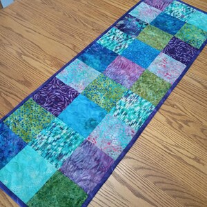 May include: A rectangular table runner featuring a patchwork design of square fabric pieces in various shades of blue, green, and purple. The runner is bordered by a solid dark purple fabric trim and displays a variety of mottled and patterned textures.