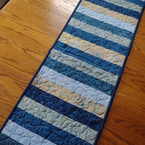 May include: A quilted table runner with a striped pattern in shades of blue, white, and yellow. The runner has a dark blue border and is placed on a wooden table. The fabric has a quilted texture.