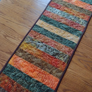 May include: A quilted table runner with a patchwork design. The runner features horizontal stripes in shades of orange, green, brown, and yellow. The edges are finished with a dark brown border. The fabric has a textured, quilted appearance.