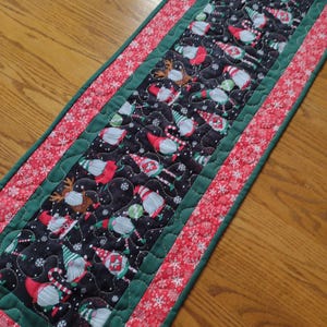May include: A quilted table runner featuring a black background with a pattern of gnomes, reindeer, and snowflakes. The runner is bordered with a red snowflake pattern and a green trim. A festive, seasonal home decor item.