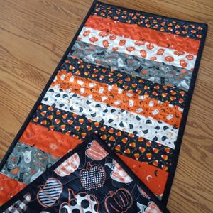May include: A rectangular quilted table runner featuring horizontal strips of fabric with Halloween patterns including orange pumpkins, white ghosts, black cats, and candy corn. The runner is finished with a black fabric border and binding.