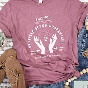 May include: A heather mauve t-shirt with a white graphic design that says "Lady M's Cleaning Company Spotless Scrub Guaranteed" with a cross and the year 1623. The text below the graphic says "What's done CAN be undone!"