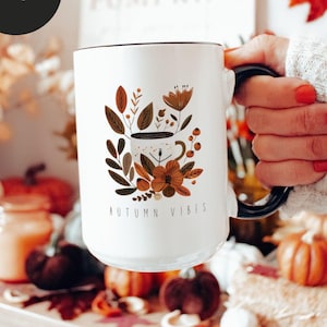 May include: White ceramic mug with a black rim and handle. The mug features a floral design with a coffee cup in the center and the text "AUTUMN VIBES".