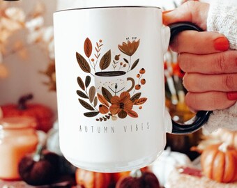 May include: White ceramic mug with a black rim and handle. The mug features a floral design with a coffee cup in the center and the text "AUTUMN VIBES".