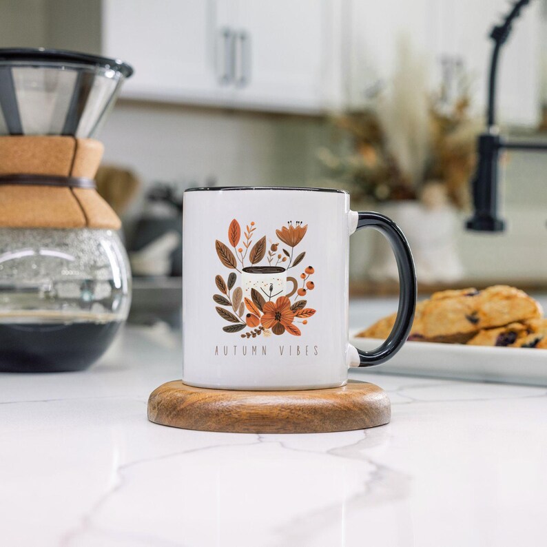 May include: White ceramic mug with a black rim and handle. The mug features a colorful illustration of a coffee cup with autumn leaves and flowers. The text "AUTUMN VIBES" is printed on the mug.