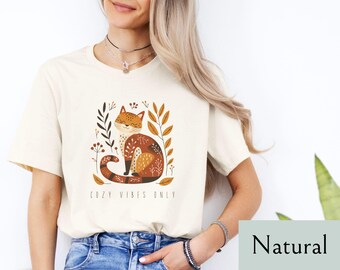 May include: A white t-shirt with a graphic of a brown and orange cat sitting in a bed of leaves. The text "Cozy Vibes Only" is printed below the cat.