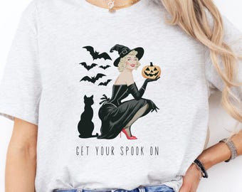 May include: A gray t-shirt with a black and white illustration of a woman in a witch costume holding a jack-o-lantern. The text "GET YOUR SPOOK ON" is printed below the illustration. The t-shirt is available in the color Ash.