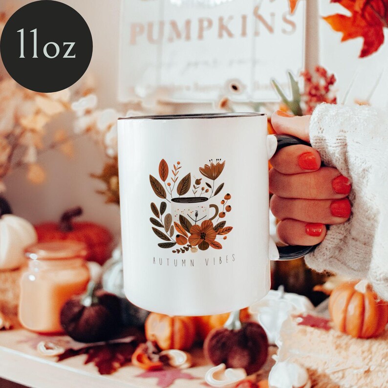 May include: White ceramic mug with a black rim and handle. The mug features a floral design with a cup of coffee and the text "Autumn Vibes".