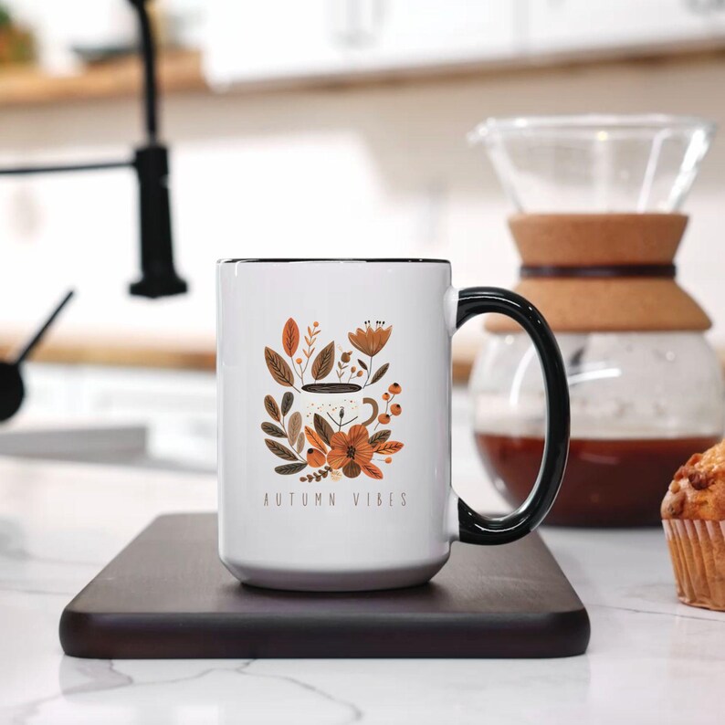 May include: White ceramic mug with a black rim and handle. The mug features a stylized illustration of a coffee cup with autumn leaves and flowers. The text "AUTUMN VIBES" is printed on the mug.