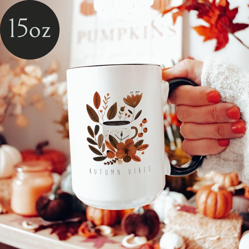 May include: White ceramic mug with a black rim and handle. The mug features a floral design with a coffee cup in the center and the text "AUTUMN VIBES".