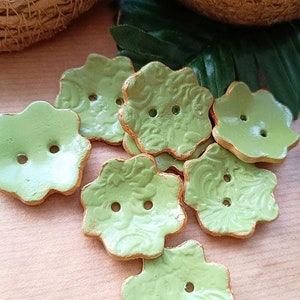 Op de afbeelding: Een set van acht groene keramische knopen met gouden rand. De knopen zijn bloemvormig met vijf bloemblaadjes en hebben elk twee gaten.