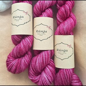 Può includere: Tre gomitoli di filato rosa e bianco con un'etichetta bianca che dice "Rünja Happy Crafting Shop".