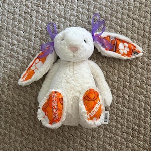 May include: A white plush bunny with orange and purple accents. The bunny has orange and white patterned ears and orange paw pads with the text "CLEMSON TIGERS". Purple ribbons are tied to the ears. A soft, cuddly stuffed animal.