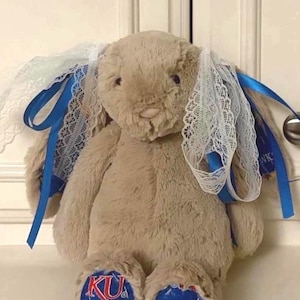 May include: A plush tan rabbit stuffed animal with long ears adorned with white lace trim and blue satin ribbons. The rabbit features blue fabric on the soles of its feet, printed with the red and blue KU logo and a yellow bird mascot.