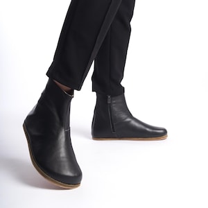 May include: Black leather ankle boots with a side zipper and a rounded toe. The boots have a low, light brown sole and are worn with black pants. The boots are a minimalist design.