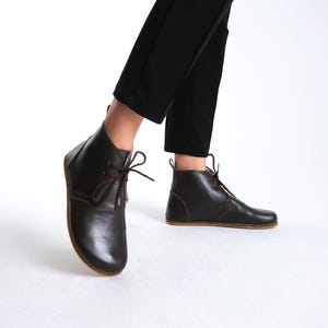May include: Dark brown leather ankle boots with brown laces. The boots have a minimalist design with a rounded toe and a flat sole. The boots are worn with black pants.