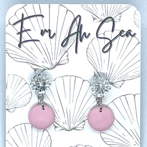 Può includere: Orecchini a disco rosa con perno color argento e un accento trasparente e scintillante. Gli orecchini sono esposti su una carta bianca con un motivo a conchiglia e il testo "Em Ah Sea" e "Handmade Clay Jewellery".