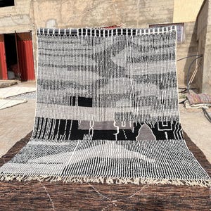 Puede incluir: Una alfombra rectangular grande con un diseño geométrico en negro, blanco y gris. La alfombra presenta rayas verticales y horizontales, junto con formas abstractas. La alfombra tiene un borde con flecos y se exhibe al aire libre.