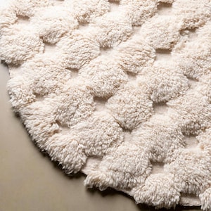 Puede incluir: Primer plano de una alfombra texturizada de color crema con un patrón repetitivo de círculos esponjosos en relieve. La alfombra tiene un aspecto suave y afelpado, lo que sugiere una textura cómoda y acogedora. El diseño es simple pero visualmente atractivo.