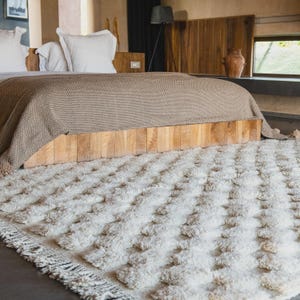 May include: A cream-colored, textured rug with a repeating pattern of raised, square tufts. The rug is placed in a bedroom setting, with a wooden bed frame and a neutral-toned blanket. The rug's edges have a fringe detail.