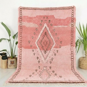 May include: A large, rectangular rug in shades of pink and brown. The rug features a central diamond pattern and a border with a fringe. The design includes geometric shapes and a textured surface, suitable for home decor.