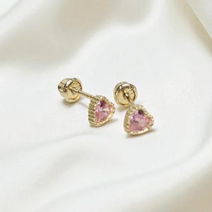 以下が含まれることがあります： ピンク色のハート型宝石が付いた、ゴールドカラーのスタッドピアス。 白いシルクのような背景に配置され、繊細なデザインと色を際立たせています。