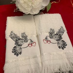 Pode incluir: Duas toalhas de mão brancas com bordas franjadas. Cada toalha apresenta um desenho bordado de um pássaro preto carregando dois corações vermelhos.
