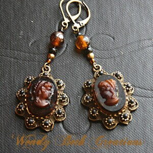 May include: A pair of gold tone earrings with cameo pendants. The cameos are brown and feature a profile of a woman's face. The earrings are accented with black beads.