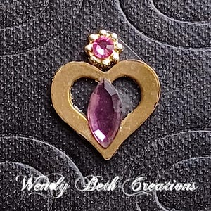 May include: A gold-colored heart-shaped charm with a purple gemstone and a small pink flower-shaped accent. The charm is set against a textured black background, with the text "Wendy Beth Creations" in cursive.