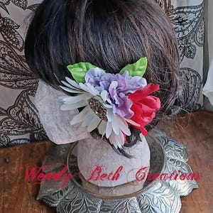 May include: A white, purple, and red flower hair clip with green leaves. The clip is on a mannequin head with dark brown hair.