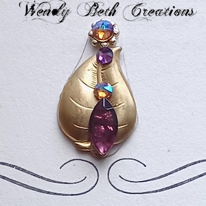 May include: A gold-toned leaf-shaped pendant with a cluster of iridescent crystals and a large, deep purple marquise-cut crystal. The pendant is adorned with the text "Wendy Beth Creations" in elegant script.