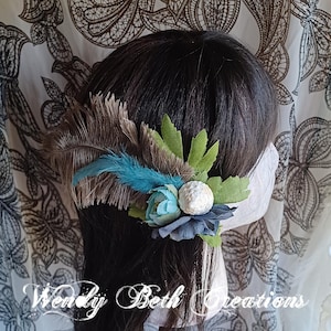 Puede incluir: Tocado decorativo con plumas marrones y turquesas, hojas verdes y flores de tela azules. Una banda blanca asegura el arreglo floral al cabello. El texto "Wendy Beth Creations" es visible en la parte inferior.