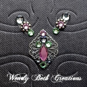 May include: A set of three decorative jewellery pieces with colourful gemstones. The set includes a central pendant with a large, purple, teardrop-shaped stone and two smaller earring-like pieces. The text "Wendy Beth Creations" is at the bottom.